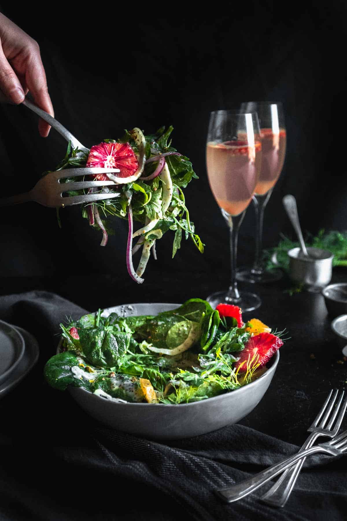 Blood orange salad being pulled from the bowl with serving utensils. The salad is next to two forks and two flutes of sparkling pink cocktails