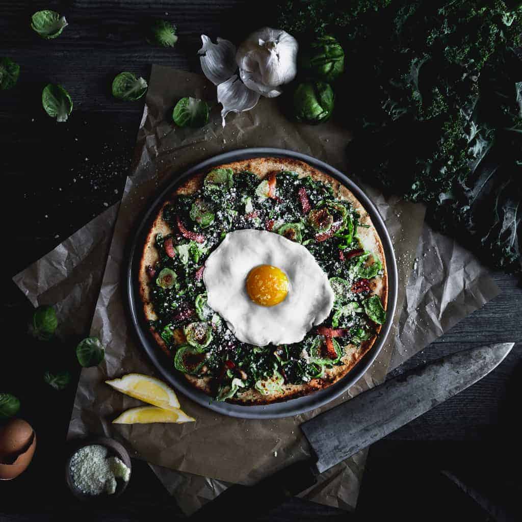 A crispy cracker crust pizza topped with shaved kale, Brussel sprouts, bacon, pecorino and a sunny side egg. The pizza is surrounded by fresh kale, garlic, Brussel sprout leaves, lemon wedges and a sharp knife.