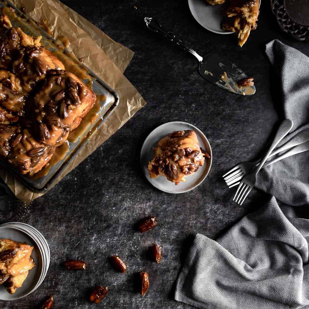 An overhead shot of a gooey Sticky Toffee Pecan Roll on a plate next to a tray of the buns, forks and a scattering of dates