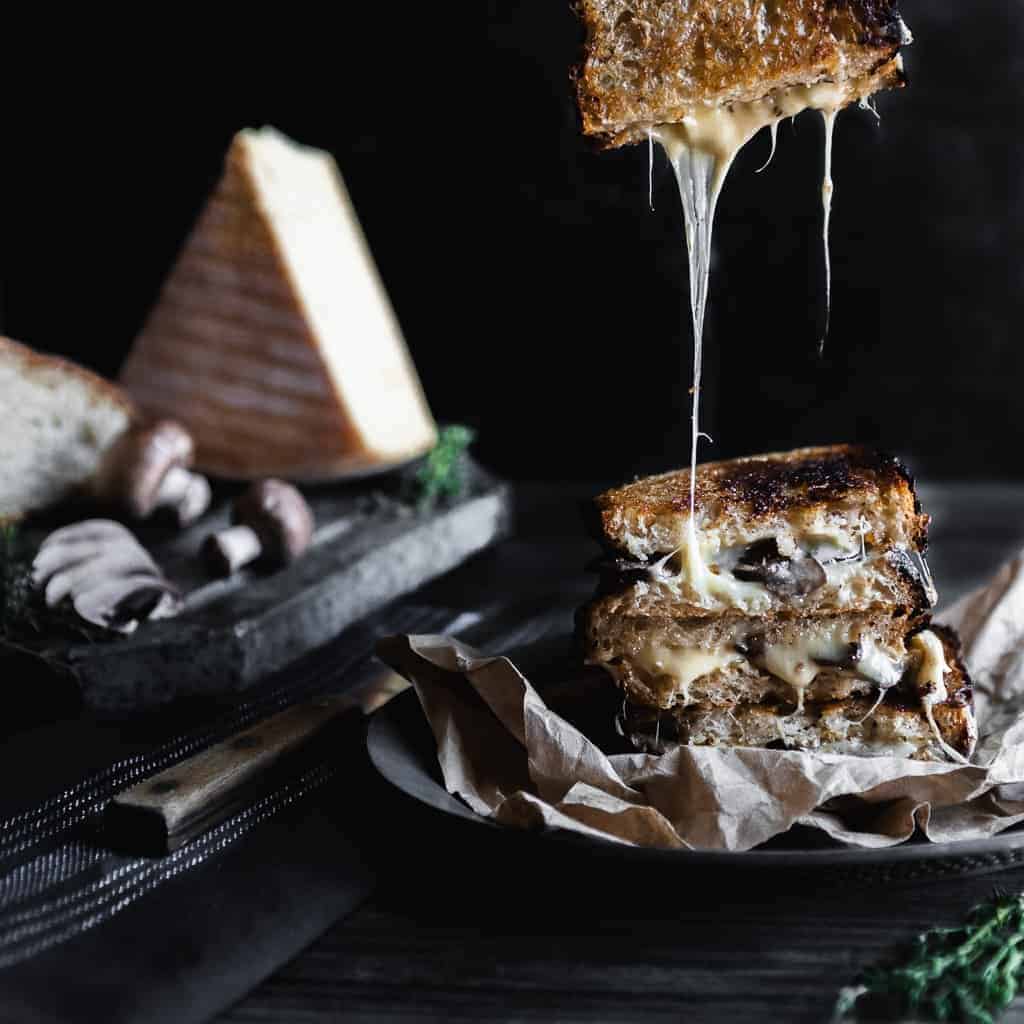 A stack of raclette grilled cheese sandwiches with one half of a sandwich being pulled up and creating a gooey cheese pull. A wedge of raclette cheese with mushrooms and a loaf of bread is in the background