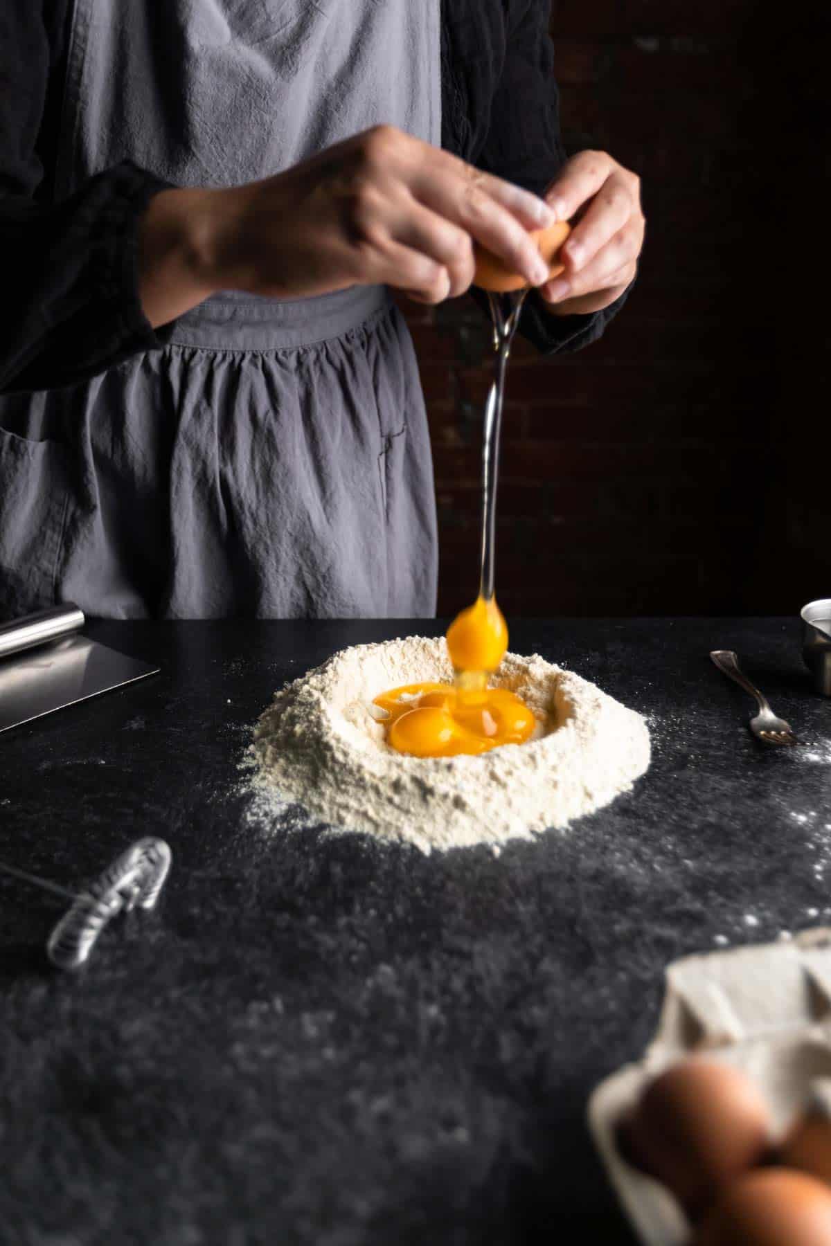 an aproned woman cracking an egg into a well of flour filled with egg yolks