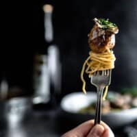 A vertical fork with a twirl of spaghetti topped with a chicken marsala meatball with a mushroom and sprinkle of parsley