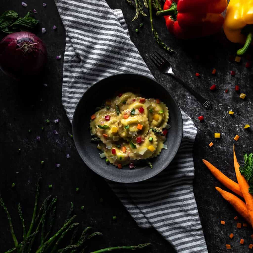 A bowl full of Funfetti Ravioli Primavera on a striped linen. The bowl is surrounded by whole and diced bell peppers, red onion, carrots and asparagus