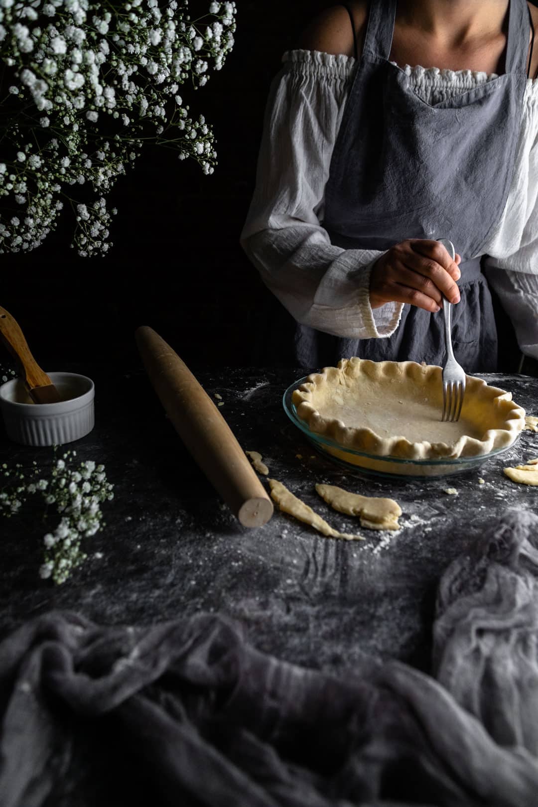 A woman using a fork to poke holes (dock) the bottom of an unbaked crimped pie crust next to a rolling pin and vase of tiny white flowers