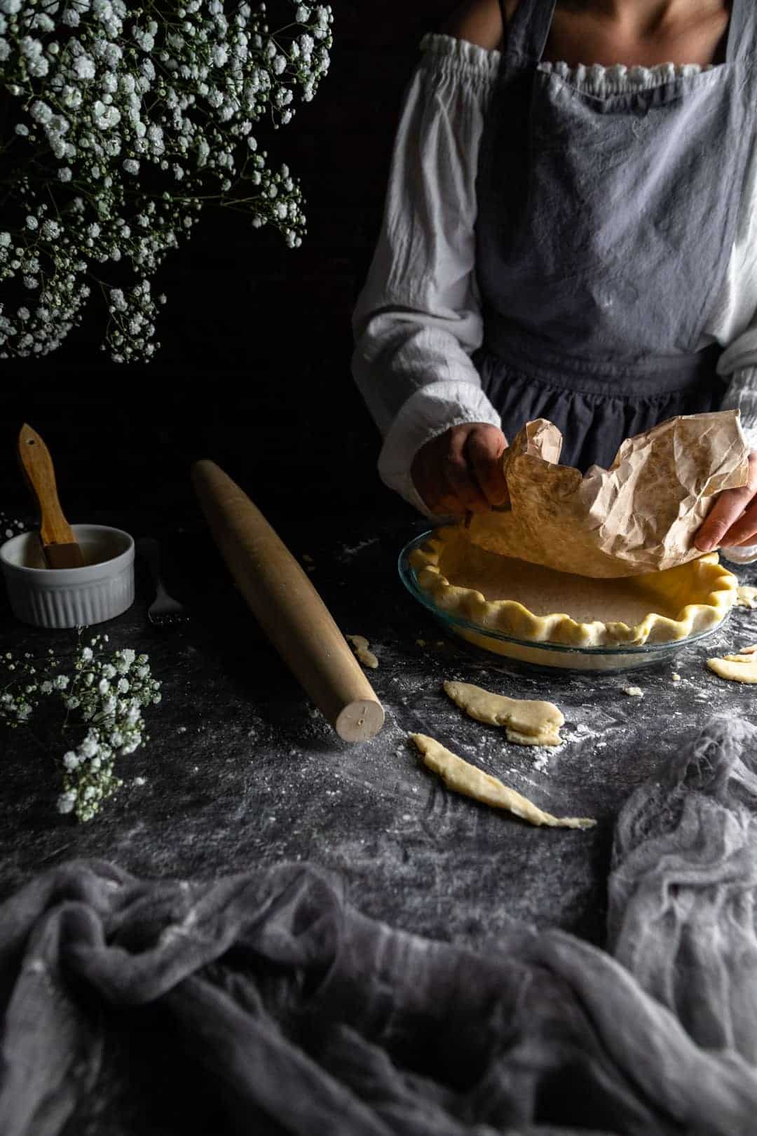 A woman placing a crumpled coffee filter into the bottom of an unbaked crimped pie crust next to a rolling pin and vase of tiny white flowers