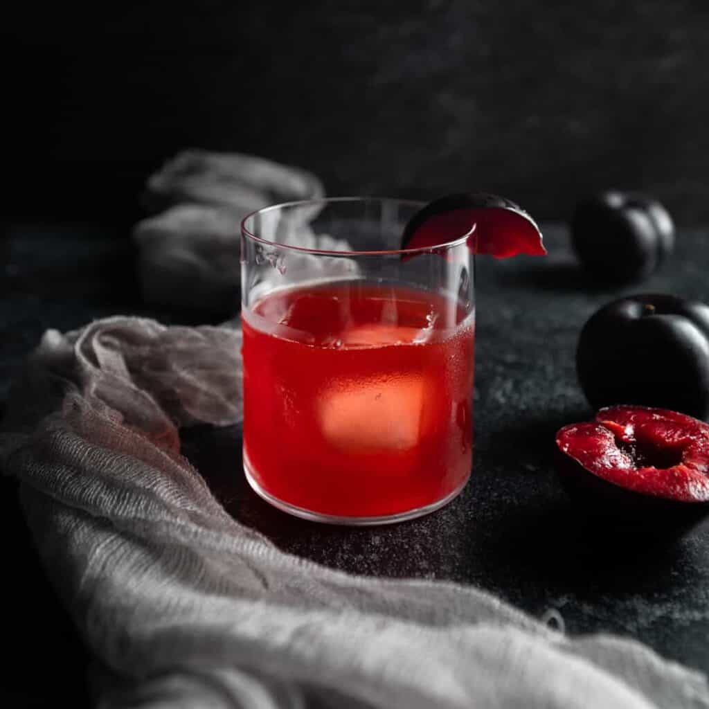 A plum rum old fashioned cocktail in a short glass on a large ice cube garnished with a slice of fresh purple plum.