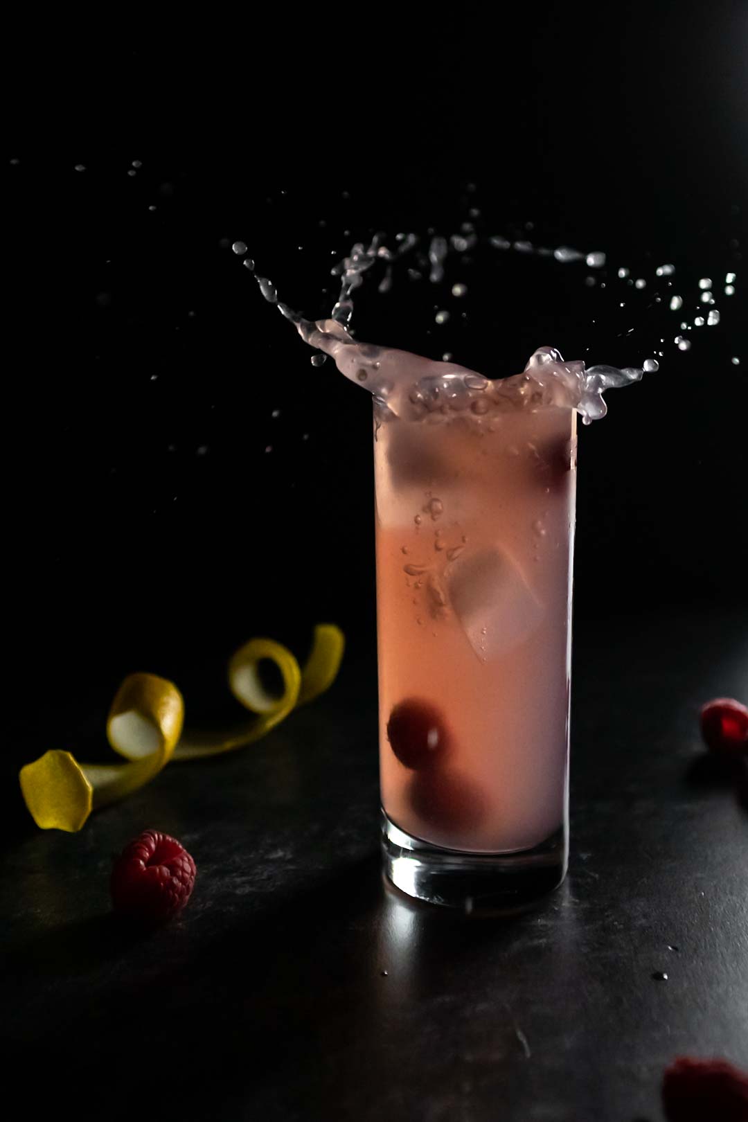 A raspberry lychee collins cocktail splashing out of a tall Collins glass. There are raspberries around the glass and a spiral of lemon peel