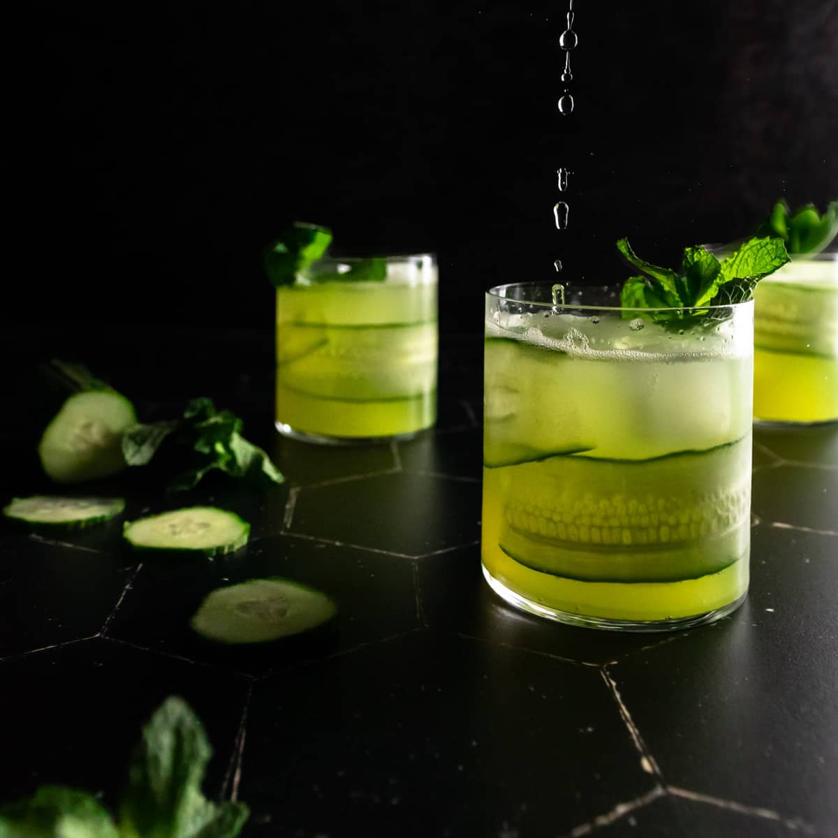 Pouring soda water into a cucumber mojito garnished with a cucumber ribbon and fresh mint. Two other cocktails and a sliced cucumber are in the background