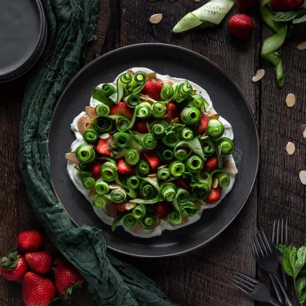 overhead view of cucumber strawberry salad. The cucumber is cut in ribbons and spiraled on a bed of goat cheese with strawberries and topped with almond slices, mint and strawberry vinaigrette. 