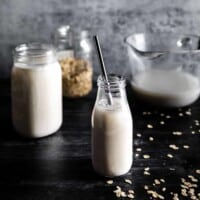 A small glass milk jar full of freshly made oat milk with a metal straw. A jar full of oat milk, a jar full of rolled oats and a mixing bowl with oat milk is in the background with a scattering of oats.