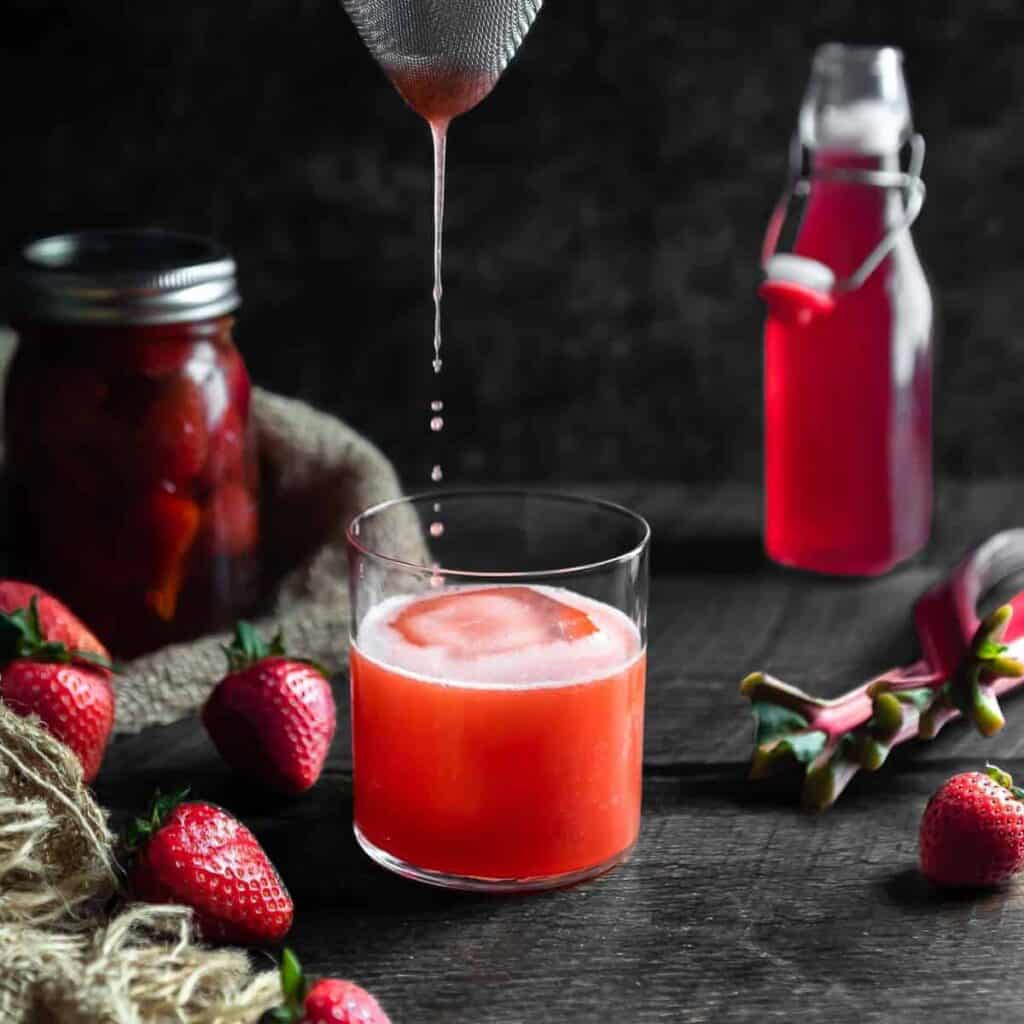 Strawberry Whiskey Sour Being poured into a glass through a strainer. The glass is surrounded by a jar of strawberry infused whiskey, fresh strawberries and a bottle of rhubarb syrup