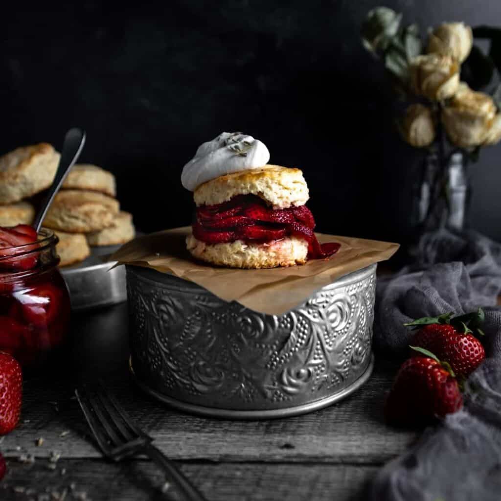 A lavender biscuit filled with lavender sugar sliced strawberries and topped with a dollop of whipped cream sitting on an upside-down tin. A jar of macerated strawberries, dried roses and a tray of lavender biscuits are in the background