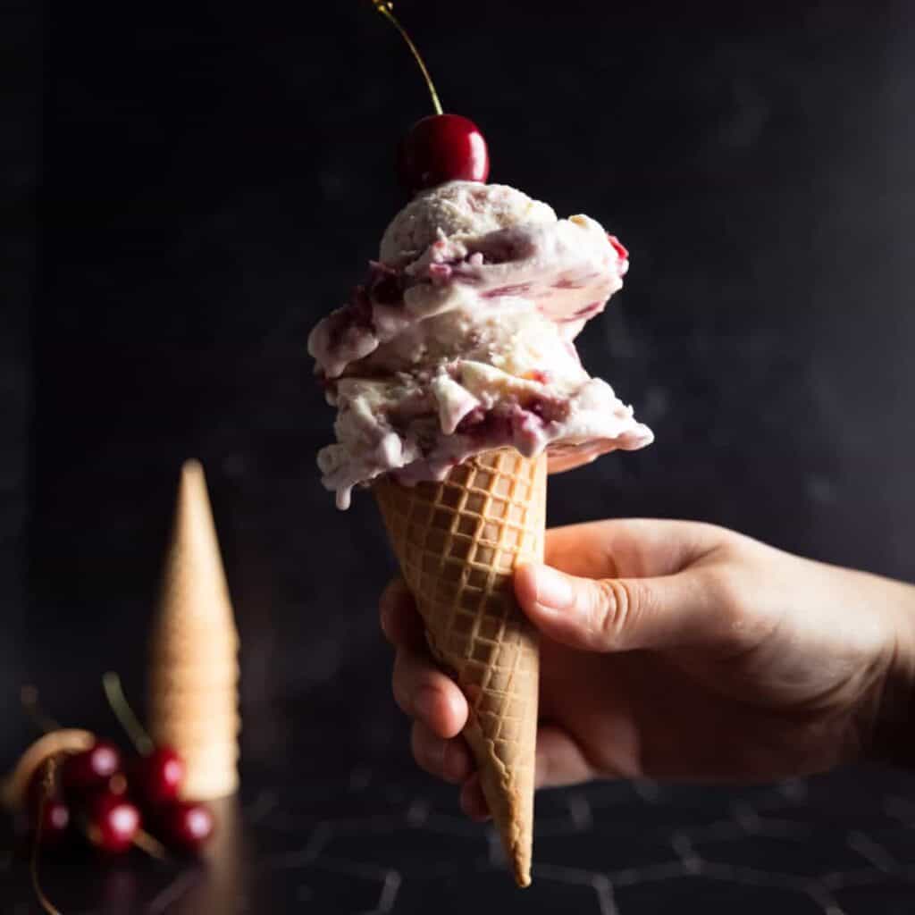 a hand holding a sugar cone topped with two scoops of Cherry Vanilla Ice Cream with a fresh red cherry on top!