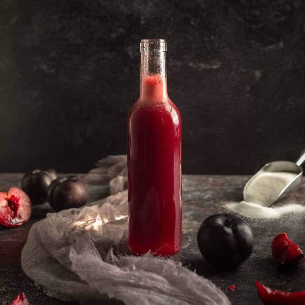 a bottle of plum syrup surrounded by whole and sliced purple plums and a scoop of granulated sugar