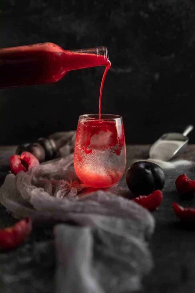 plum syrup being poured into a stemless wine glass filled with ice and soda water. the glass is surrounded by fresh sliced plums and a scoop of sugar