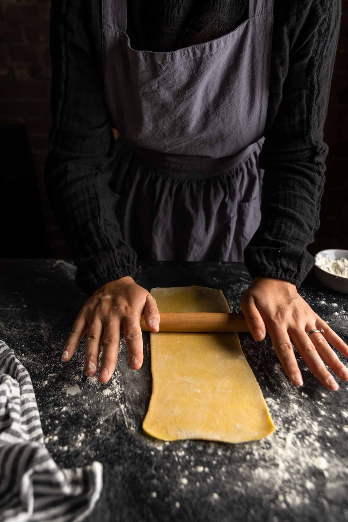 a woman rolling a long thin sheet of semolina pasta dough to make fettuccine