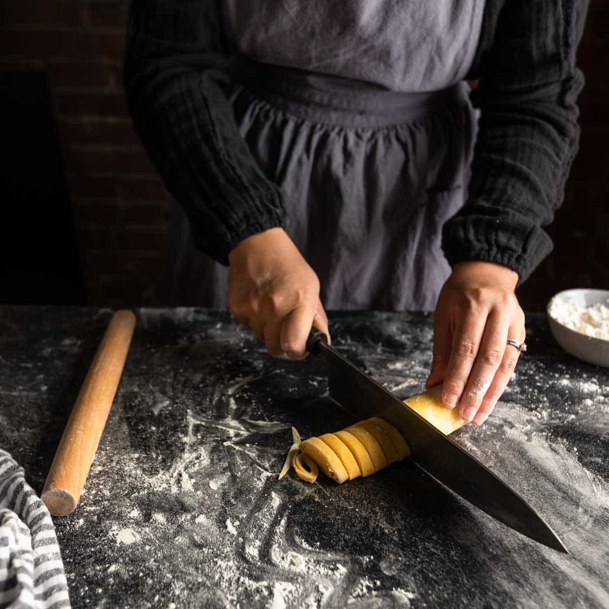a woman using a sharp knife to cut a log of rolled pasta dough into spirals of homemade fettuccine pasta