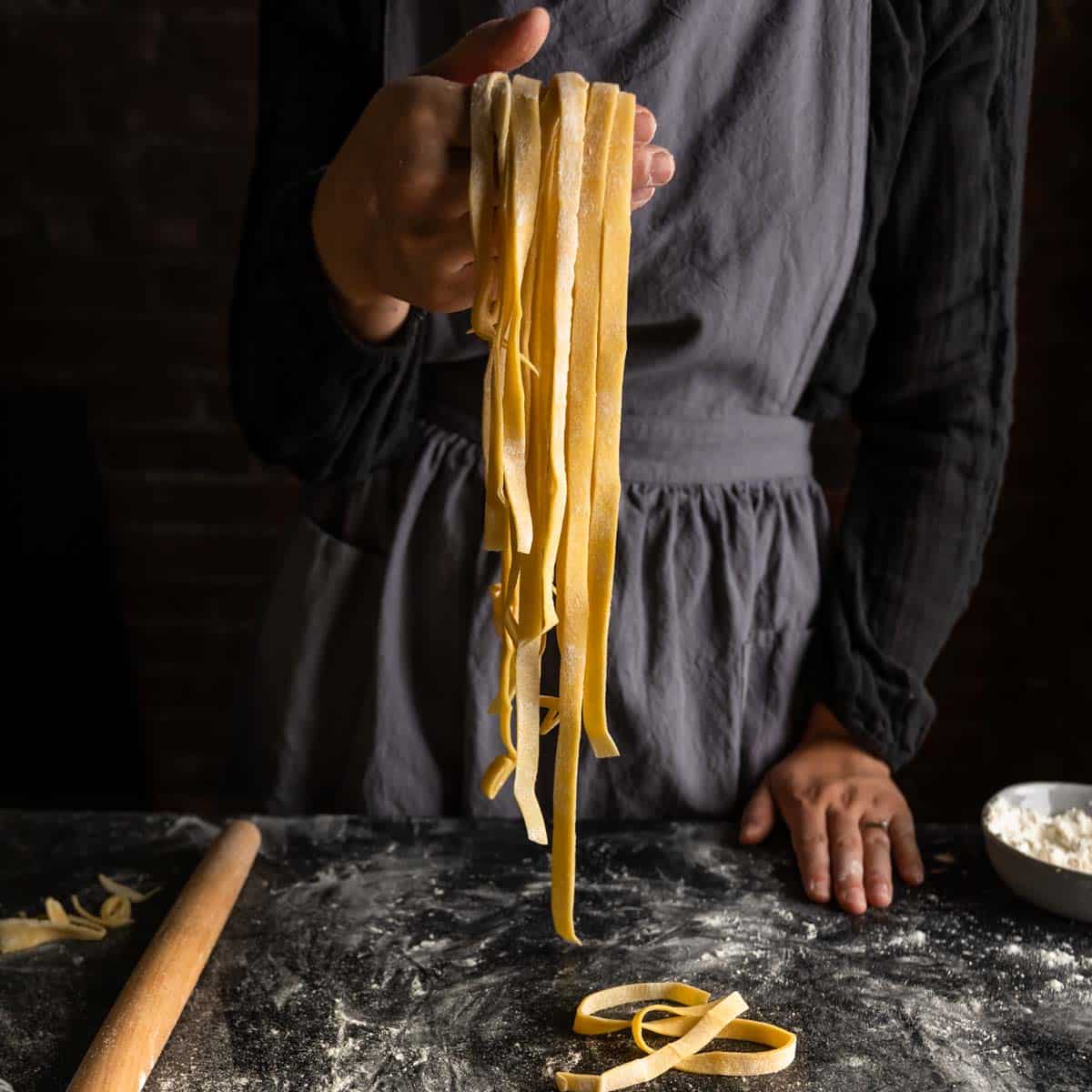 An aproned woman holding up a handful of homemade fettuccine pasta noodles
