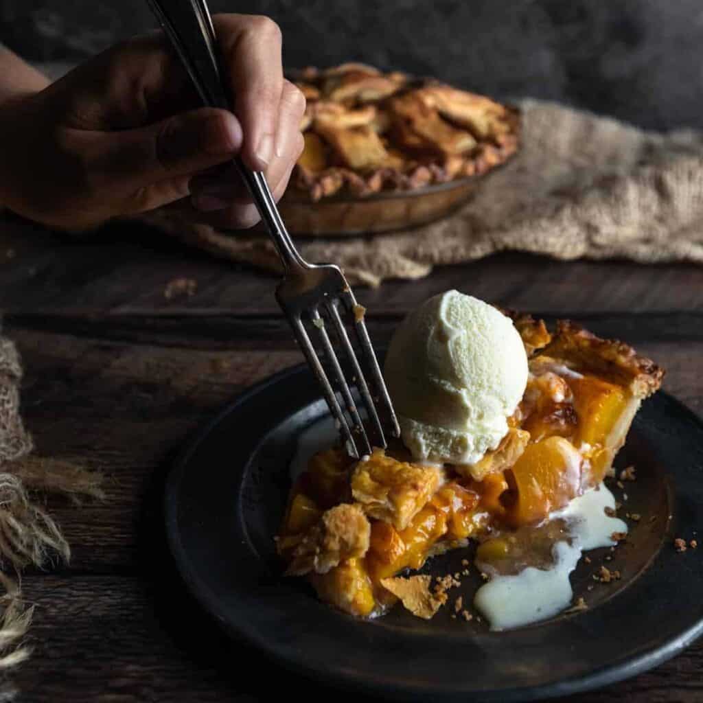 A fork going into a big slice of peach mango pie topped with vanilla ice cream