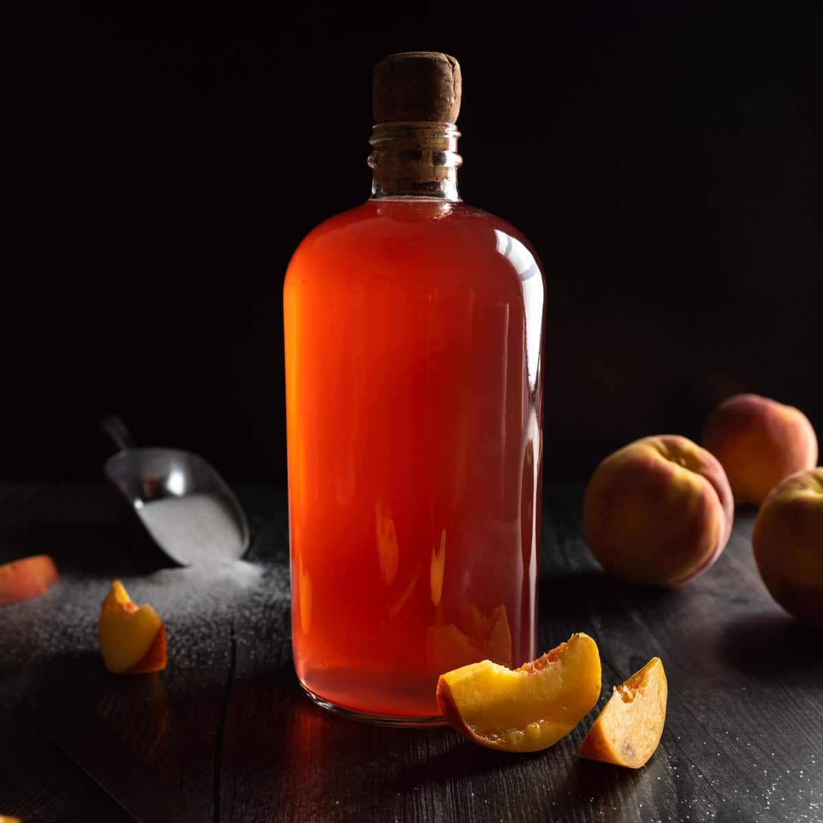 A corked glass bottle of peach syrup surrounded by whole and sliced peaches and a scoop or sugar