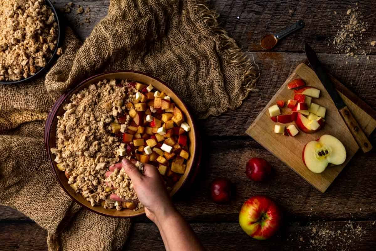 A hand sprinkling the crumble topping over the plum and apple filling next to a cutting board with diced apple.