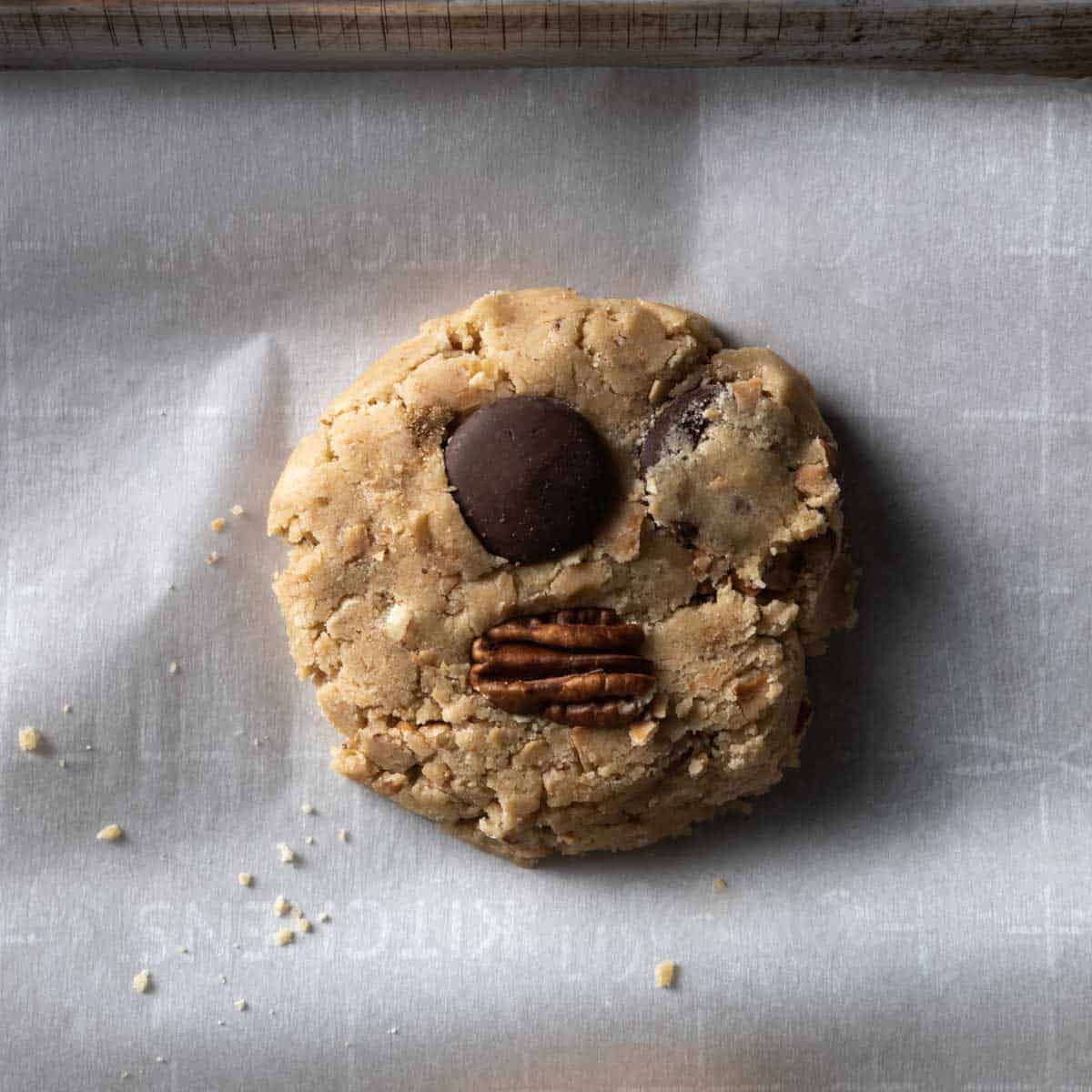 A slightly flattened scoop of brown butter cookie dough on a sheet tray topped with a dark chocolate disk and toasted pecan half 