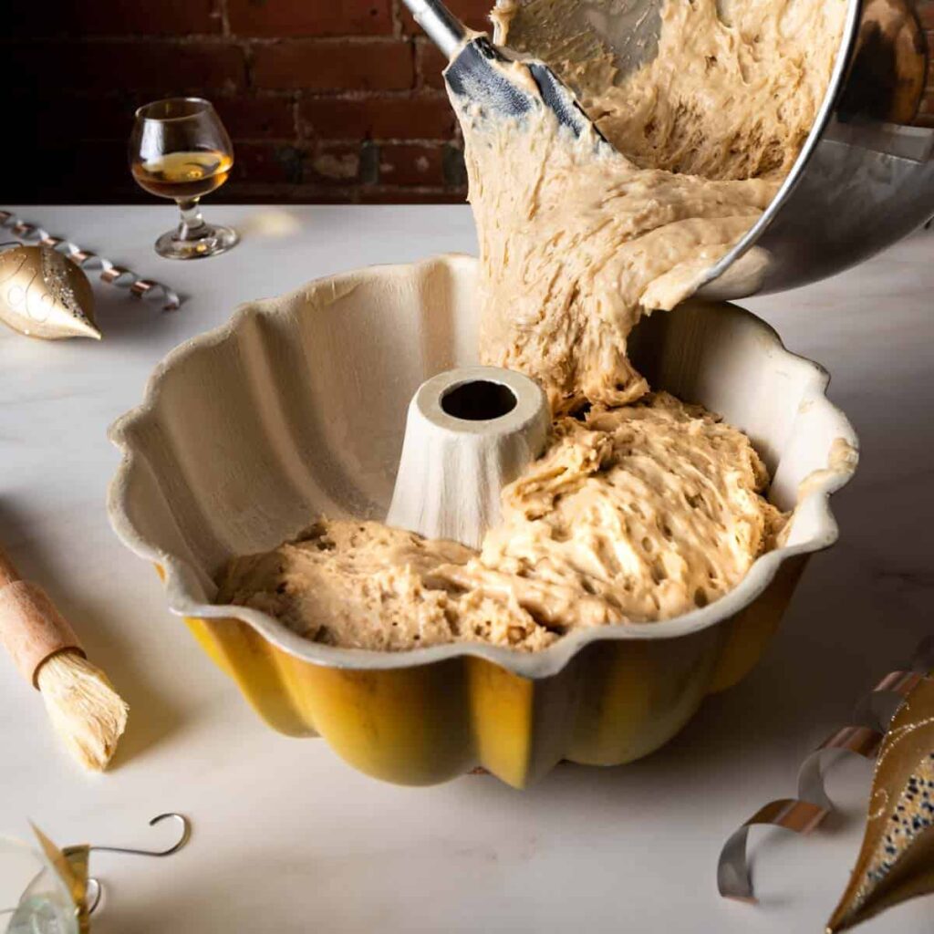 Pouring thick eggnog cake batter from a mixing bowl into a prepared bundt pan