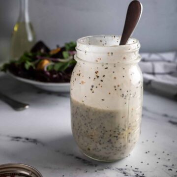 An open mason jar of homemade poppyseed dressing next to a green salad and a sprinkle of poppy seeds