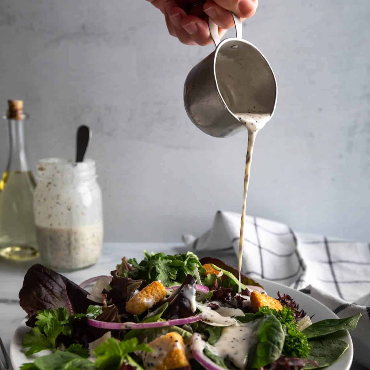 A small tin of creamy poppyseed dressing being poured over a green salad