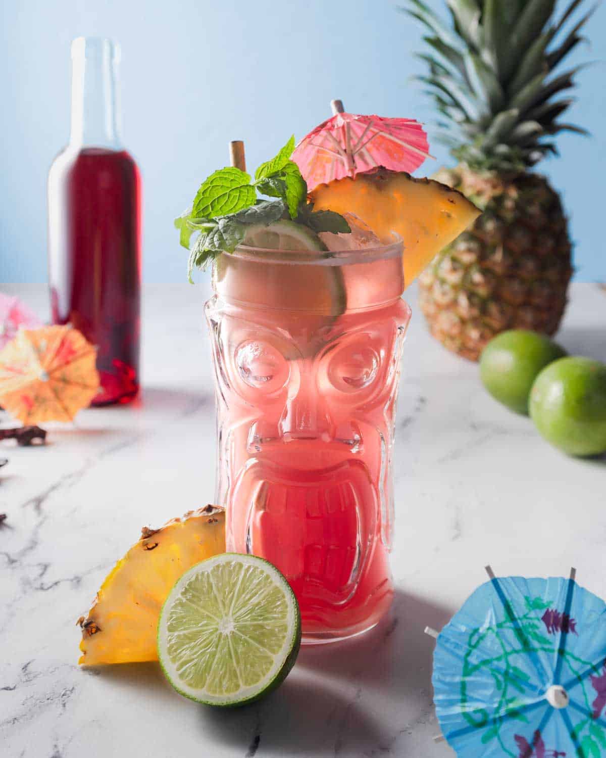 A tall tiki glass of hibiscus rum punch garnished with pineapple, lime, mint and cocktail umbrellas