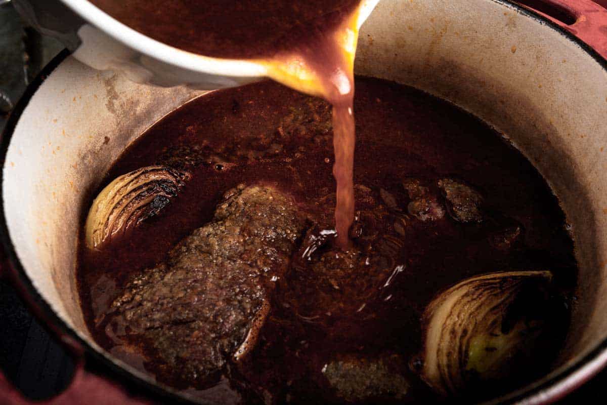 Korean flavored braising liquid being poured over short ribs and onion in a dutch oven pot
