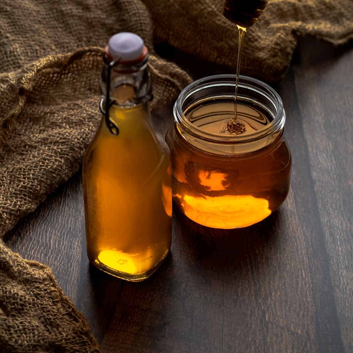 A bottle of honey syrup and honey dripping into a jar