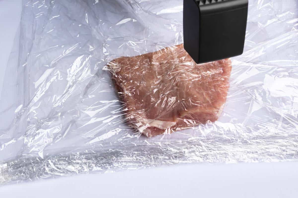 A piece of pork wrapped in plastic wrap being pounded with a mallet