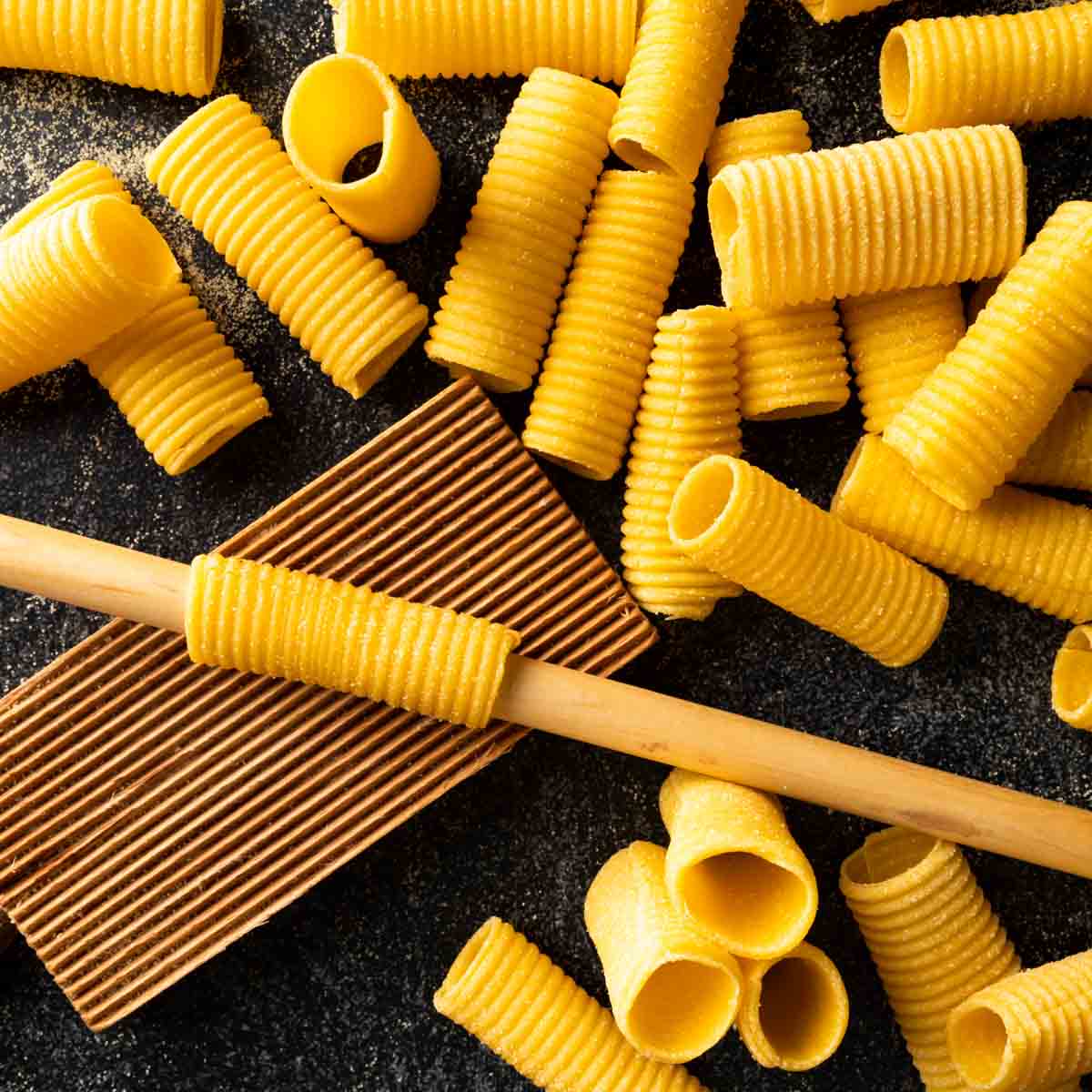 A picture of several homemade rigatoni and one being made with a dowel and gnocchi board