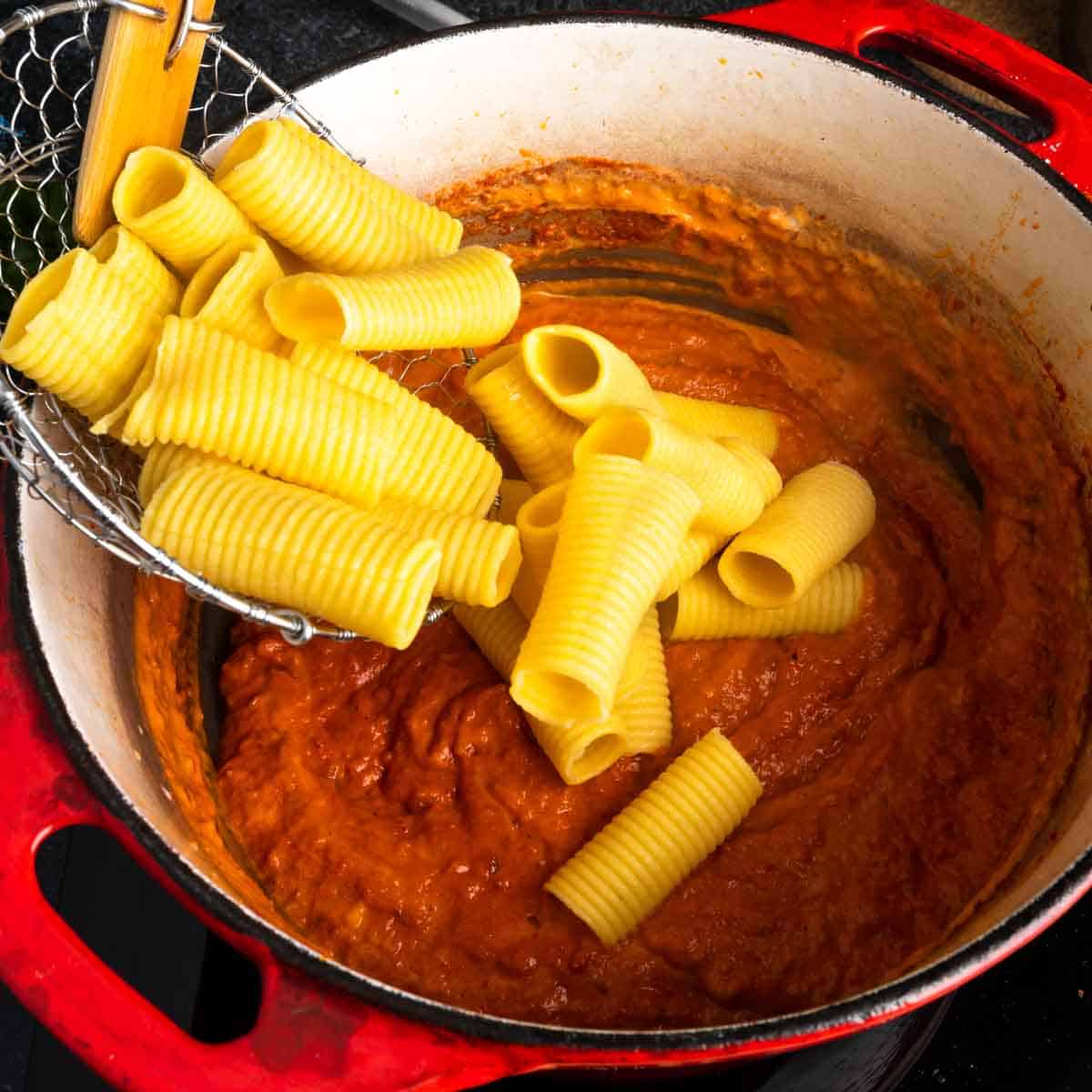 Dumping boiled rigatoni into a big pot of sauce