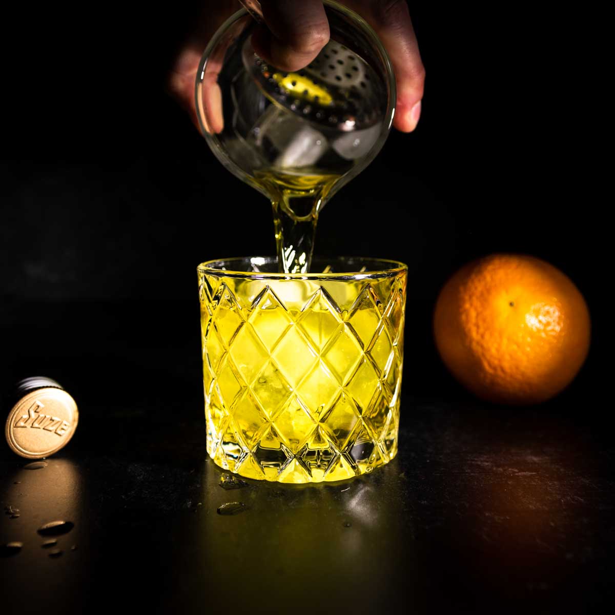 Straining a white negroni over a sphere of ice in an old fashioned glass