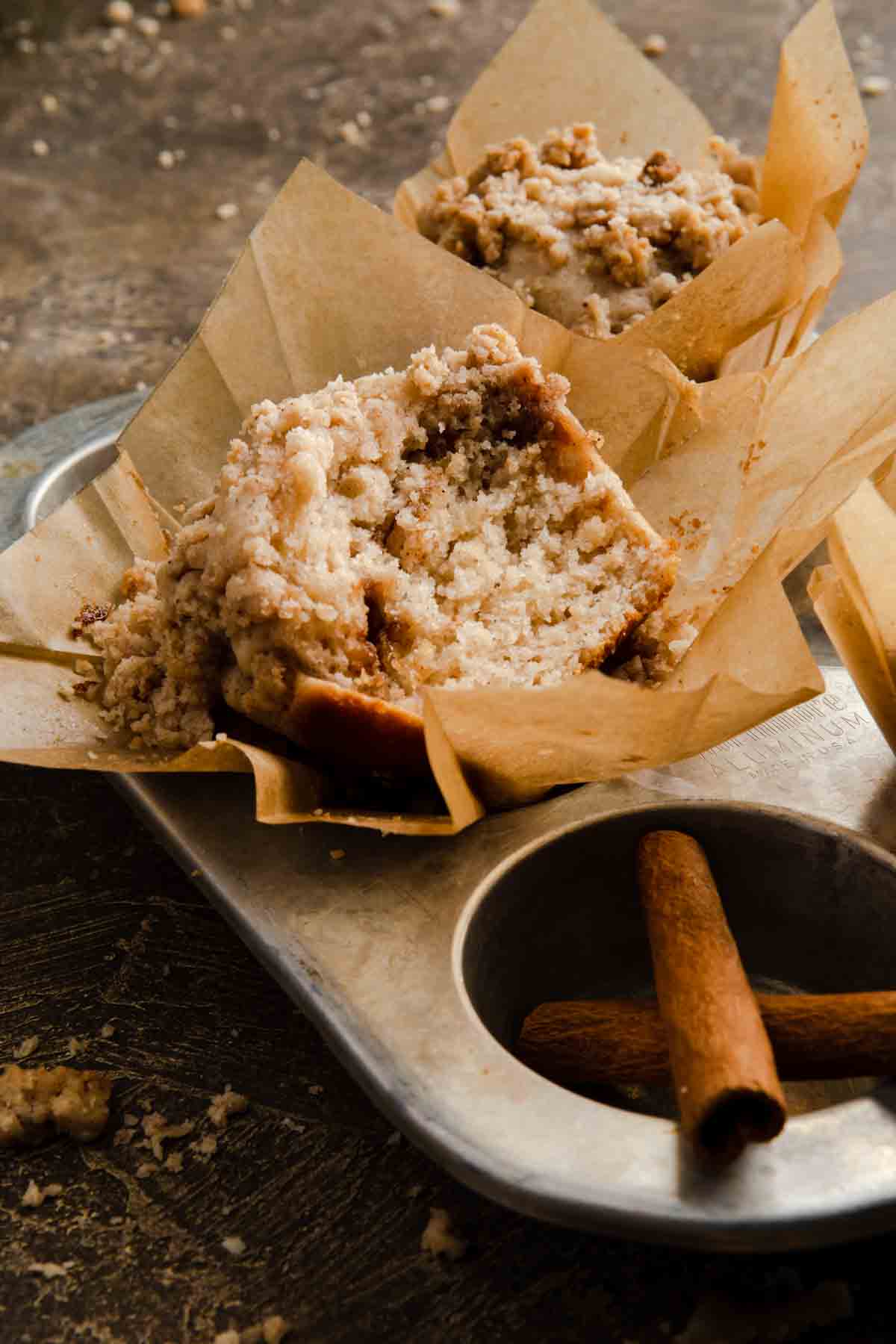 A cinnamon crumb muffin in a muffin tin with cinnamon sticks