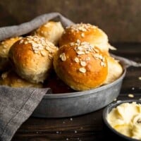 A tin filled with fluffy golden oatmeal rolls and aside of soft butter