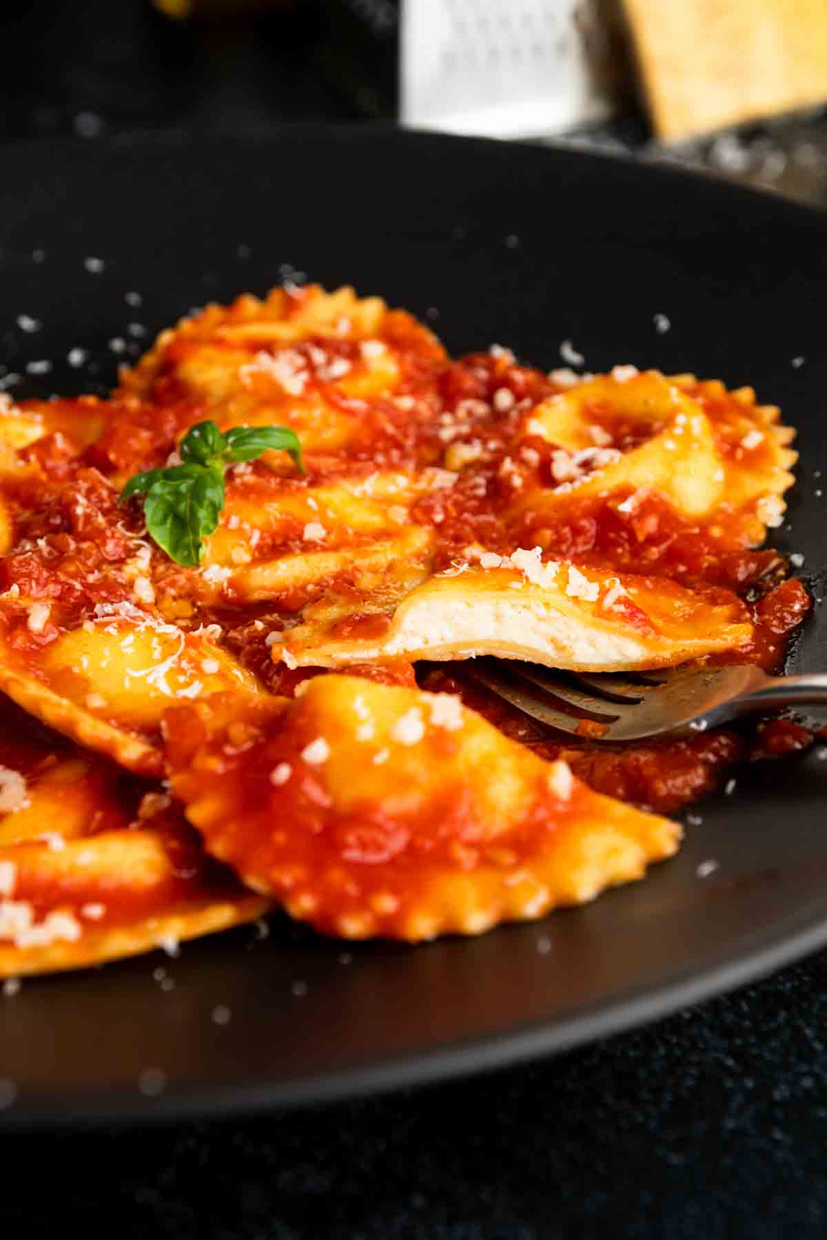 A fork with half of a homemade cheese ravioli on plate of ravioli and Italian red sauce. 