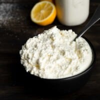 A bowl of homemade whole milk ricotta cheese next to a jar of cream and a halved lemon.