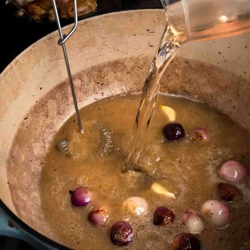 Whisking chicken stock and white wine into the Dutch oven with pearl onions and garlic.